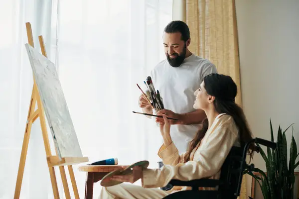 Good looking bearded man watching disabled beautiful wife on wheelchair painting on easel at home — Stock Photo