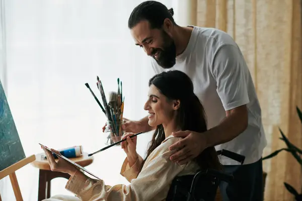 Soins joyeux mari aider son inclusive belle femme à peindre sur chevalet tout en à la maison — Photo de stock
