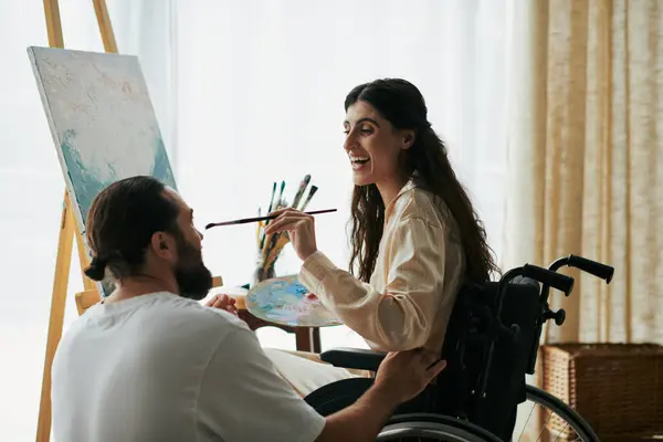 Позитивний чоловік допомагає своїй інклюзивній красивій дружині на інвалідному візку малювати на мольберті вдома — стокове фото