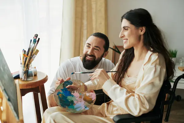 Positiver Ehemann hilft seiner inklusiven schönen Frau im Rollstuhl, zu Hause auf Staffelei zu malen — Stockfoto