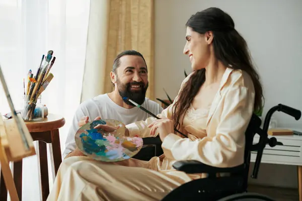 Marito positivo che aiuta la sua bella moglie inclusiva sulla sedia a rotelle a dipingere sul cavalletto mentre a casa — Foto stock