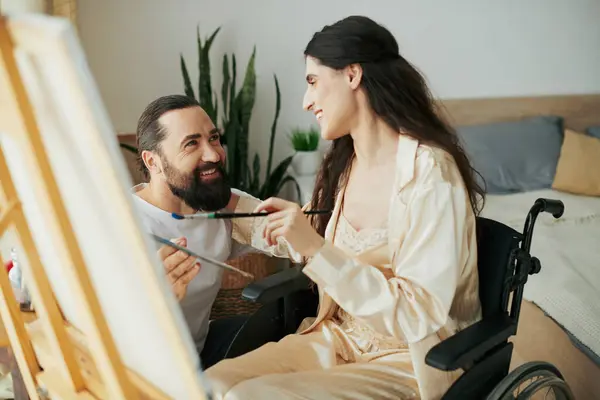 Attraktive Frau mit Gehbehinderung malt auf Staffelei neben ihrem fröhlichen bärtigen Mann — Stockfoto