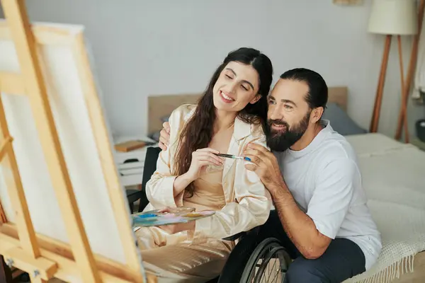 Attraente donna con disabilità motoria pittura su cavalletto accanto al marito barbuto allegro — Foto stock