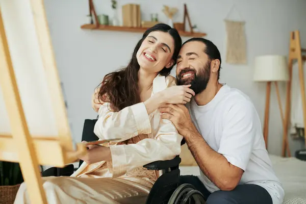 Mulher atraente com mobilidade deficiência pintura no cavalete ao lado de seu marido barbudo alegre — Fotografia de Stock