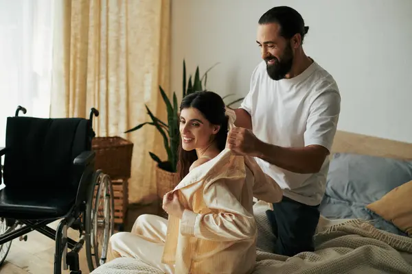 Весёлый любящий мужчина в домашней одежде помогает своей жене-инвалиду переодеться в пижаму в спальне дома — стоковое фото