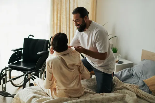Красивый любящий мужчина в домашней одежде помогает своей жене-инвалиду переодеться в пижаму в спальне дома — стоковое фото