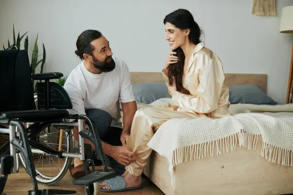 Homem amoroso barbudo em roupa de casa ajudando sua esposa deficiente a se transformar em pijama no quarto em casa — Fotografia de Stock