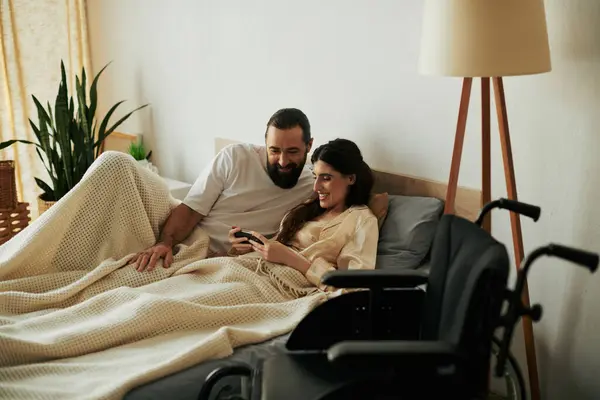 Freudige Frau mit Gehbehinderung, die mit Handy neben ihrem bärtigen, liebevollen Ehemann im Bett liegt — Stockfoto