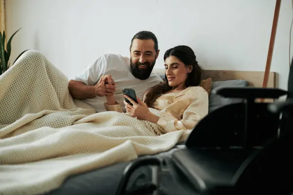 Mulher alegre com deficiência de mobilidade deitada na cama com telefone ao lado de seu marido amoroso barbudo — Fotografia de Stock