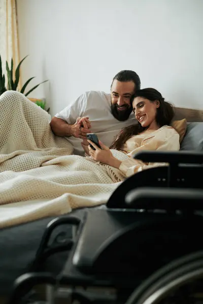 Mujer alegre con discapacidad de movilidad acostada en la cama con teléfono al lado de su marido barbudo amoroso - foto de stock
