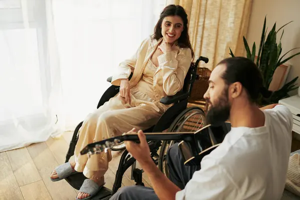 Donna allegra con disabilità in sedia a rotelle a guardare il marito barbuto suonare la chitarra per lei — Foto stock