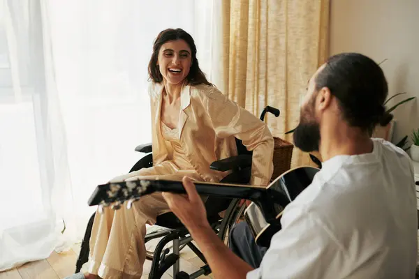 Веселая женщина с инвалидностью в инвалидном кресле наблюдает, как ее бородатый муж играет на гитаре для нее — стоковое фото