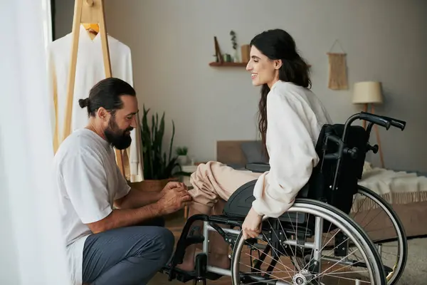 Barbudo hombre amoroso ayudando a su esposa inclusiva en silla de ruedas a vestirse mientras está en el dormitorio - foto de stock
