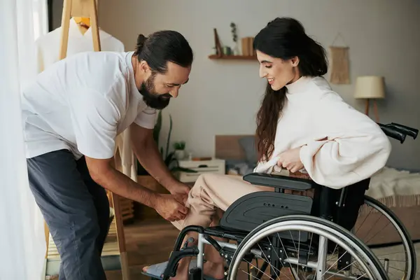 Barbuto amorevole uomo aiutare la sua moglie inclusiva sulla sedia a rotelle per vestirsi mentre in camera da letto — Foto stock