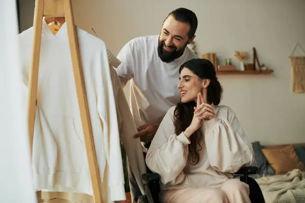 Barbuto uomo premuroso aiutando la sua moglie inclusiva sulla sedia a rotelle per vestirsi mentre in camera da letto — Foto stock