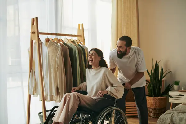 Guapo hombre cariñoso ayudando a su mujer inclusiva en silla de ruedas a vestirse mientras está en el dormitorio - foto de stock