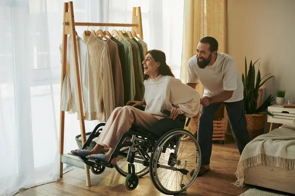 Homem carinhoso bonito ajudando sua esposa inclusiva na cadeira de rodas para se vestir enquanto no quarto — Fotografia de Stock