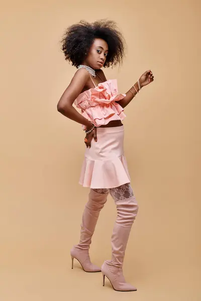 Jovem menina afro-americana na década de 20 posando em top pêssego ruffle e botas sobre o joelho em pano de fundo bege — Fotografia de Stock
