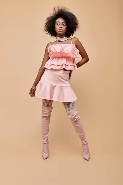 Fancy african american woman in 20s, posing in peach ruffle top and over-knee boots on beige — Stock Photo