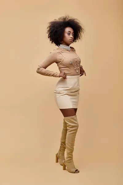 Ragazza afro-americana alla moda in mini gonna pastello e stivali alti su uno sfondo beige — Foto stock