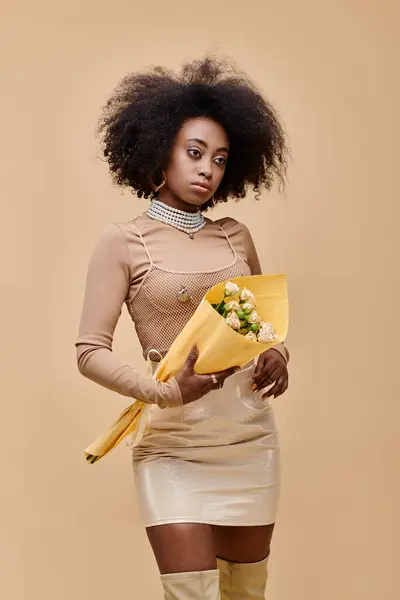 Young african american model posing in pastel peach outfit and holding flowers on beige backdrop — Stock Photo