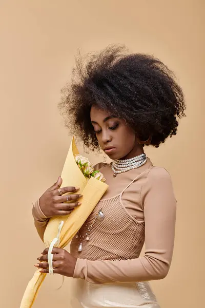 Гарна афро-американська модель позує в пастельному персиковому вбранні і тримає квіти на бежевому тлі — стокове фото