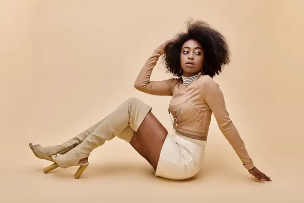 Chica afroamericana en traje de moda de pelusa de melocotón y botas altas de muslo se reclina sobre un fondo beige - foto de stock