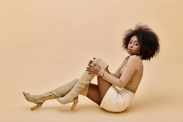 Chica americana africana rizada en traje de moda de pelusa de melocotón y botas altas de muslo se reclina en un beige - foto de stock