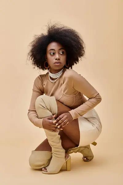 Encaracolado Africano americano mulher na moda pêssego fuzz roupa e coxa-altas botas sentado em um bege — Fotografia de Stock