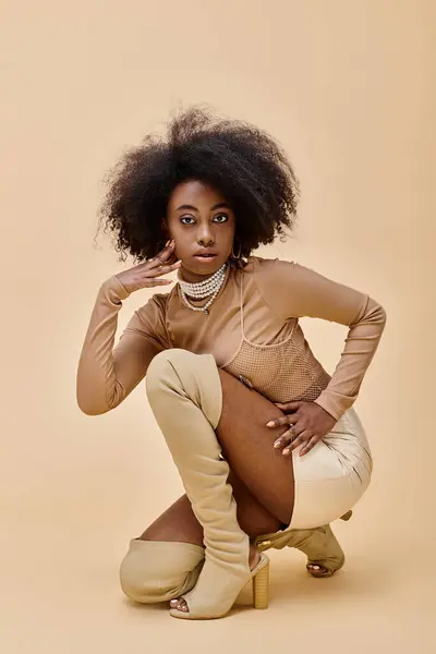 Ricci modello afroamericano in abito alla moda color pesca fuzz e stivali alti seduti su un beige — Foto stock