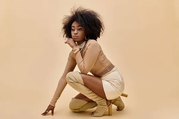 Мрійлива афро-американська модель в стильному вбранні і черевиках на стегнах позує на бежевому фоні — стокове фото
