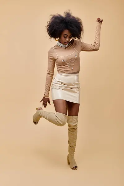African american model in trendy outfit and thigh-high boots standing on one leg on beige backdrop — Stock Photo