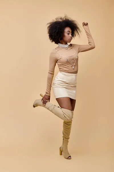 African american woman in trendy outfit and thigh-high boots standing on one leg on beige backdrop — Stock Photo