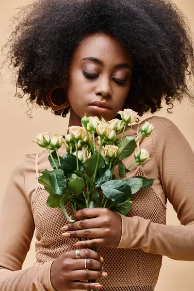 Young and curly african american woman looking at tiny roses on beige background, peach fuzz color — Stock Photo