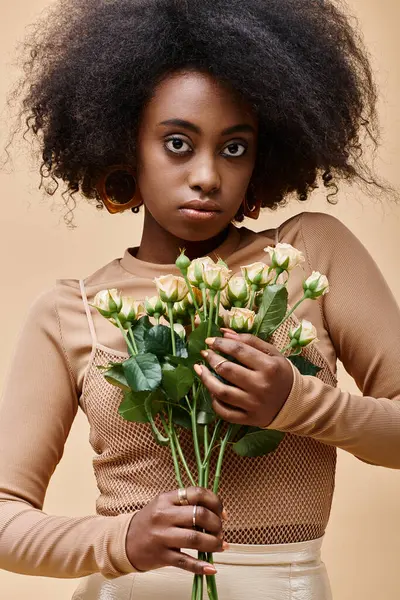 Young curly african american woman holding tiny roses on beige background, peach fuzz color — Stock Photo
