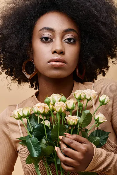 Молодая и кудрявая африканская американская девушка в возрасте 20 лет держит крошечные розы на бежевом фоне, персиковый пушок — стоковое фото