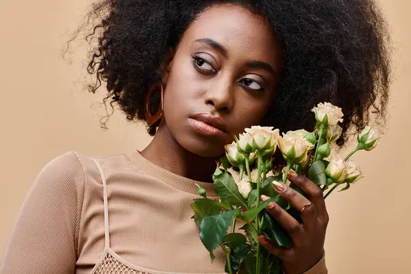 Pensive curly african american woman holding tiny roses on beige background, peach fuzz color — Stock Photo