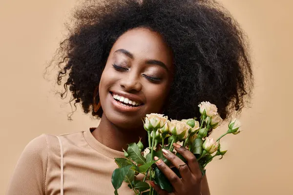 Joyeuse femme afro-américaine frisée tenant de minuscules roses sur fond beige, couleur pêche fuzz — Photo de stock