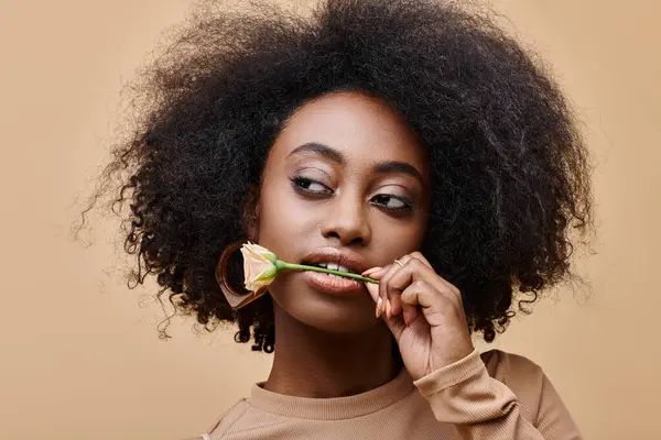 Young and curly african american woman with small rose in teeth on beige background, peach fuzz — Stock Photo