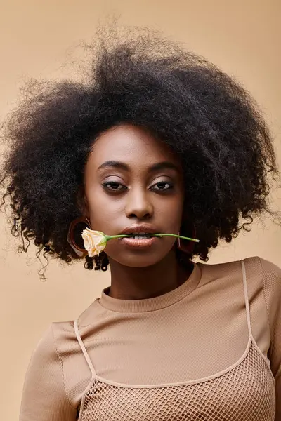 Young curly african american woman with small rose in teeth on beige background, peach fuzz — Stock Photo