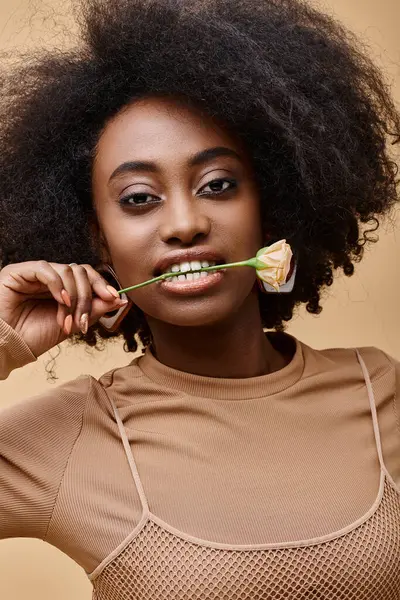 Daring curly african american woman in 20s biting small rose on beige background, peach fuzz color — Stock Photo