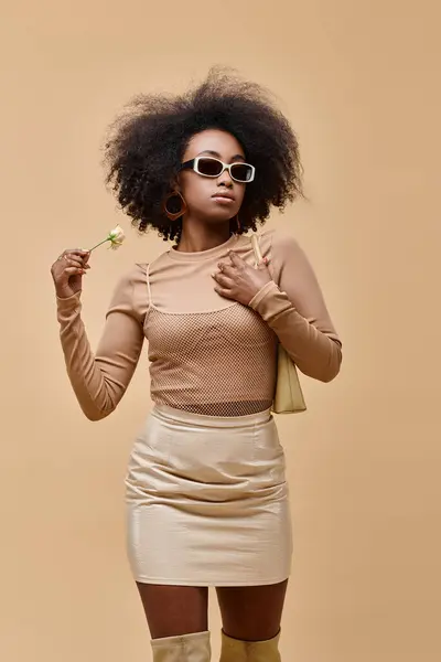 Elegante donna afroamericana in occhiali da sole in possesso di borsetta e piccola rosa su sfondo beige — Foto stock