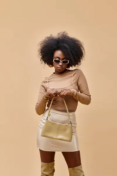 Mujer afroamericana joven y elegante en gafas de sol con bolso de moda sobre fondo beige - foto de stock