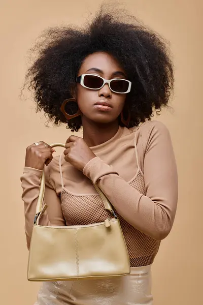 Молода і стильна афроамериканка в сонцезахисних окулярах тримає модну сумку на бежевому фоні — стокове фото