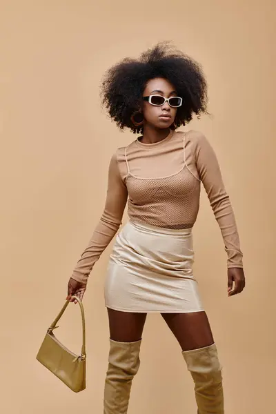 Giovane ed elegante ragazza afroamericana in occhiali da sole in possesso di borsa alla moda su sfondo beige — Foto stock