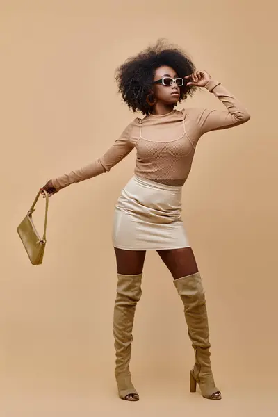 Mujer afroamericana en gafas de sol y botas de muslo alto sosteniendo bolso sobre fondo beige - foto de stock