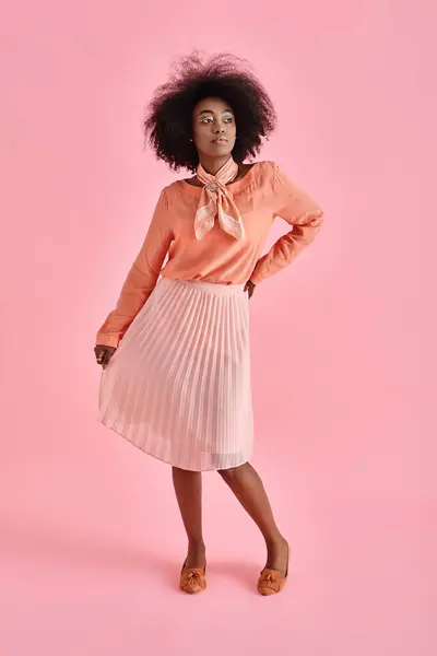 Елегантна афроамериканка в персиковій блузці і спідниці міді позує на пастельному рожевому фоні — стокове фото