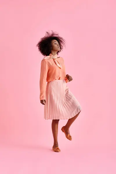 Кучерява афроамериканська жінка в персиковій блузці і спідниці міді позує на пастельному рожевому фоні — стокове фото