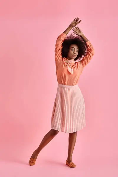 Mujer americana africana rizada en blusa de melocotón y falda midi posando con la mano levantada sobre rosa pastel - foto de stock