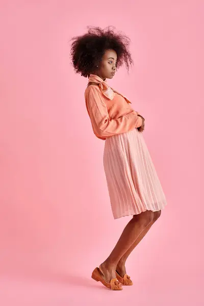 Femme afro-américaine bouclée en chemisier pêche et jupe midi regardant loin sur fond rose pastel — Photo de stock